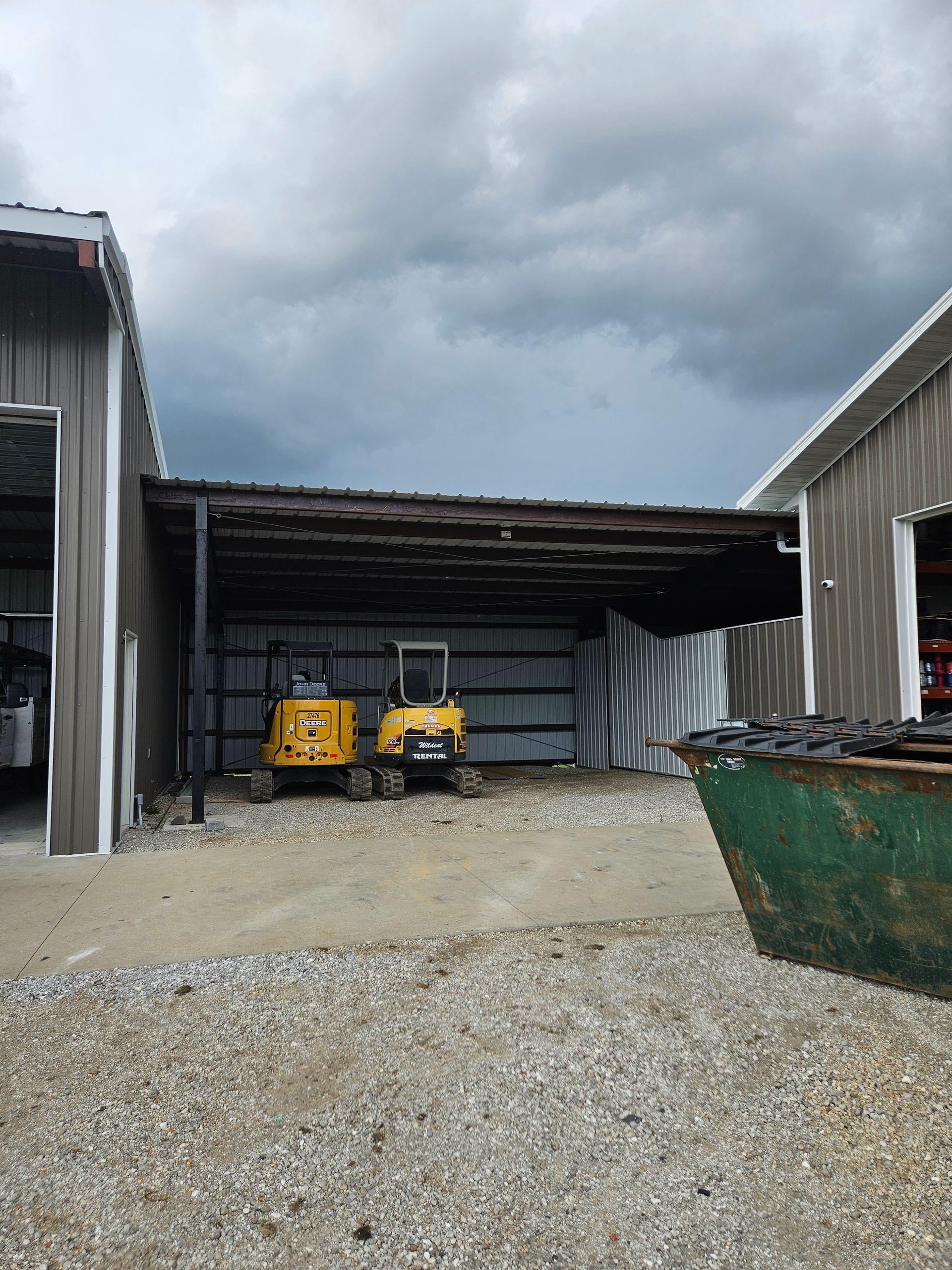 Two yellow excavators parked under a weathered awning between two metal buildings, gravel ground. Overcast sky.